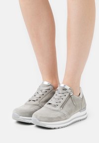 Jalat memakai sepatu olahraga suede abu-abu muda dengan sol putih, depan tali, resleting samping, dan aksen perak metalik di atas latar belakang putih.