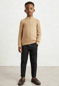 Pull en maille torsadée beige avec col montant, pantalon noir ajusté et mocassins marron. Les vêtements présentent un design texturé et une coupe ajustée.