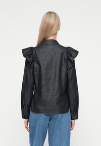 Svart metallisk bluse med rynkede skuldre, lange ermer og knappedetaljer. Kombinert med lys blå denimjeans som viser en avslappet passform.