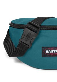 Teal tygväska med ett svart justerbart rem och spänne. Har dragkedjestängning och en logotyp patch i röd och vit på en svart bakgrund.