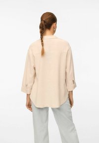 Chemise en lin beige à coupe décontractée, manches longues retroussées au niveau des poignets et ourlet arrondi. Texture lisse et drapé subtil.
