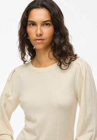 Pull en tricot beige avec de longues manches bouffantes et un col rond, présentant une texture lisse et une silhouette ajustée.