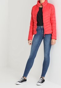 Rød quiltet jakke med høj krave og lynlås, båret over en sort skjorte. Kombineret med stramme blå jeans og sorte sneakers.