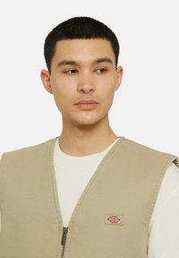 Beige arbeidsvest med glidelås foran, laget av slitesterk canvas. Har en logopatch på venstre bryst og brede, åpne armhull.