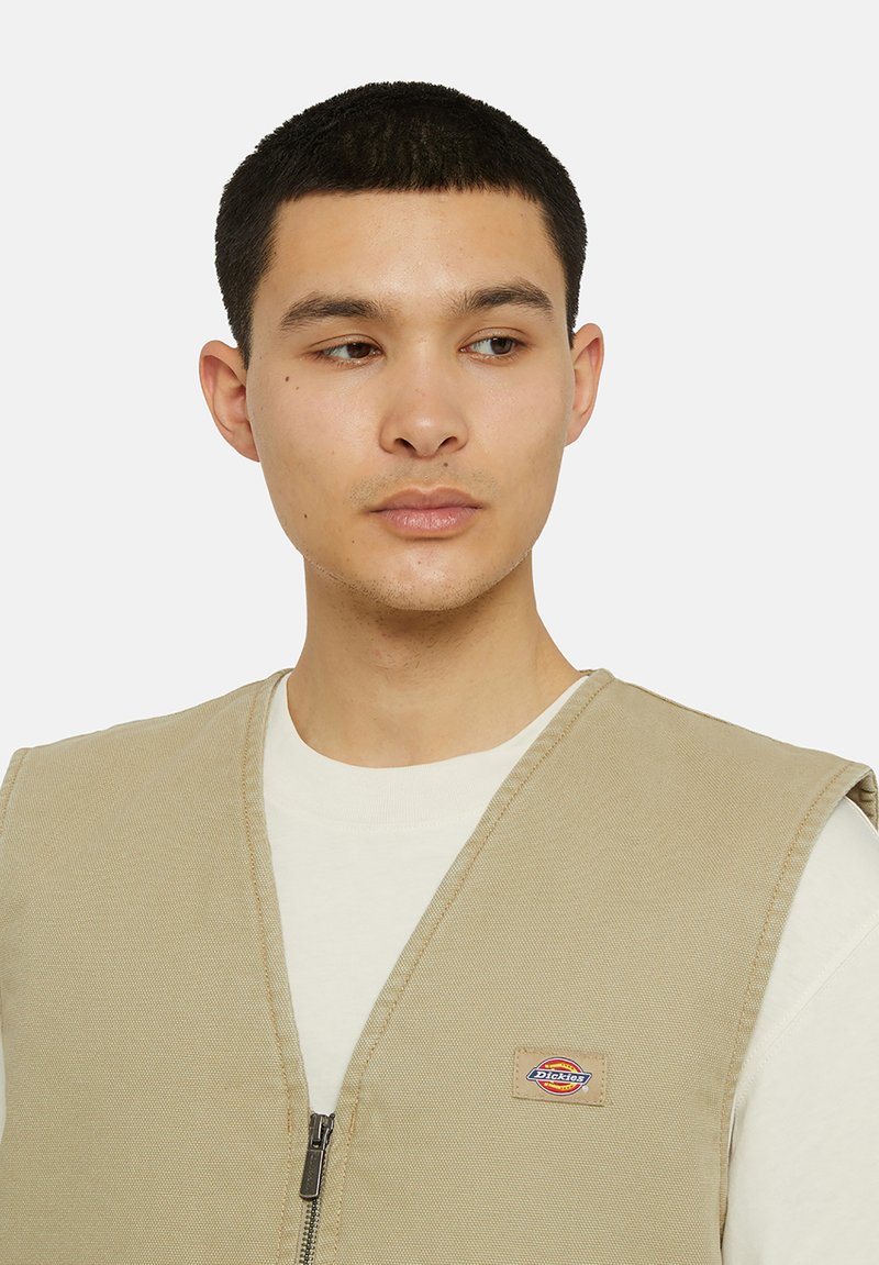 Beige Arbeitsweste mit Reißverschluss vorne, aus strapazierfähigem Canvas. Mit einem Logo-Patch auf der linken Brust und weiten, offenen Armausschnitten.