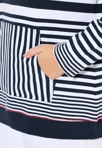 Pull en rayures avec des bandes horizontales noires et blanches. Présente une poche avant et une couture rouge en accent au bas. Texture de tissu douce.
