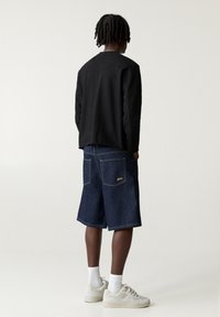Chemise noire à manches longues, short en denim avec poches à l'avant et à l'arrière, et baskets beiges. Des chaussettes blanches montantes sont visibles.