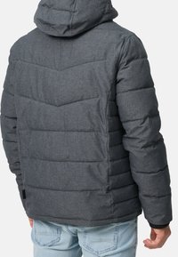 Man som bär en mörkgrå quiltad jacka med huva och ljusblå jeans, stående med ryggen mot kameran.