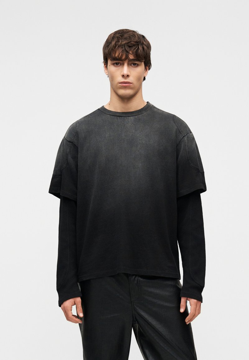 Νεαρός άνδρας που φοράει στρωμένα μαύρα μακρυμάνικα πουκάμισα κάτω από κοντομάνικο μαύρο ξεθωριασμένο t-shirt και μαύρο υφασμάτινο παντελόνι, στέκεται μπροστά σε λευκό φόντο.