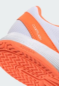 Hvid atletiksko med klare orange accenter. Har en tekstureret mesh-overdel og en gummiydersål med et tydeligt slidmønster. "COURTFLASH" og "ADIWEAR" mærker er synlige.