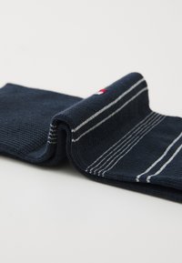 Marineblaue Socke aus gewebtem Stoff, mit weißen Streifen am oberen Bund und einem kleinen Logo in Rot und Weiß nahe dem Bund.