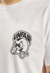 Camiseta de algodón blanca con un gráfico negro central de un personaje con una sonrisa pícara, ojos prominentes y puños, con el texto "INJUSTO" encima.