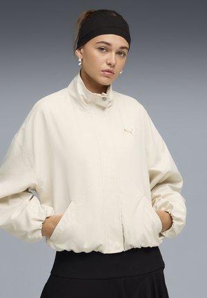 Mujer con las manos en los bolsillos, lleva una chaqueta crema con cremallera, una diadema negra y pendientes plateados contra un fondo gris degradado.