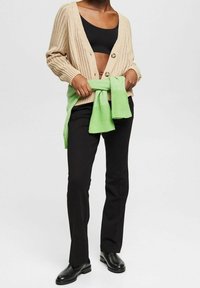 Beige gerippte Strickjacke mit Holzknöpfen, kombiniert mit einem schwarzen Crop-Top, schwarzen Hosen und einem grünen Strickpullover, der in der Taille gebunden ist.