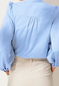 Blusa azzurra chiara con maniche a sbuffo e schiena plissettata. Realizzata in materiale morbido, presenta un colletto classico e una vestibilità rilassata.