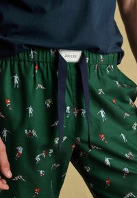 Zaļi kokvilnas šorti ar sporta tēmas apdruku, uz kuriem attēloti dažādi spēlētāji. Šorti ir ar elastīgu jostasvietu un tumši zilu pievilkšanas auklu.