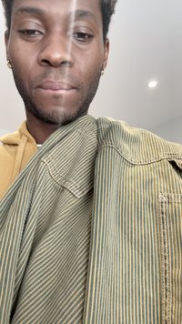 Tecido listrado em verde e amarelo pendurado, mostrando uma textura suave e costura visível. Um capuz mostarda em contraste visível por baixo.
