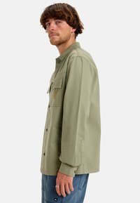 Olive green button-up shirt with a collar, two chest pockets, and a textured fabric. Features a small logo on the left pocket.