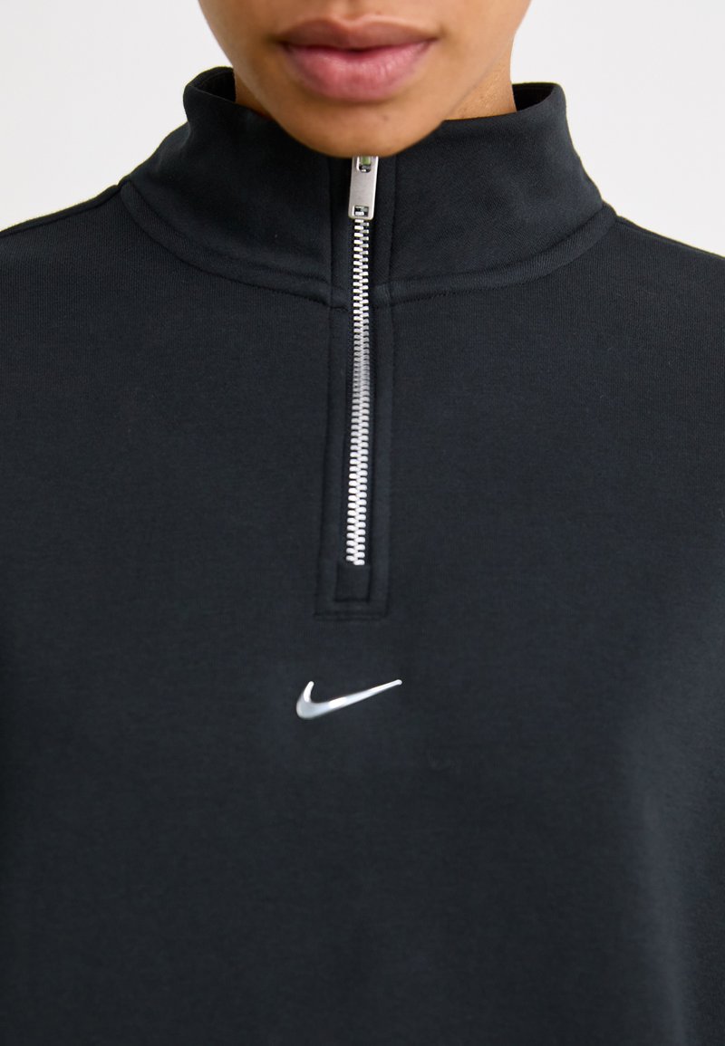 Top deportivo negro con cierre, de cuello alto, fabricado en tejido suave; presenta un logo plateado de Nike en el pecho y un detalle de cremallera plateado.