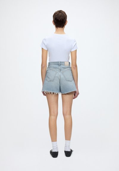 Personne debout de dos, portant un haut blanc à manches courtes, un short en jean bleu clair taille haute, des chaussettes blanches et des chaussures noires.