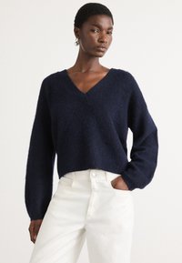 Navyblauwe, korte trui van zacht, aaibaar materiaal met een V-hals en lange, losse mouwen, gecombineerd met witte, high-waisted broek.