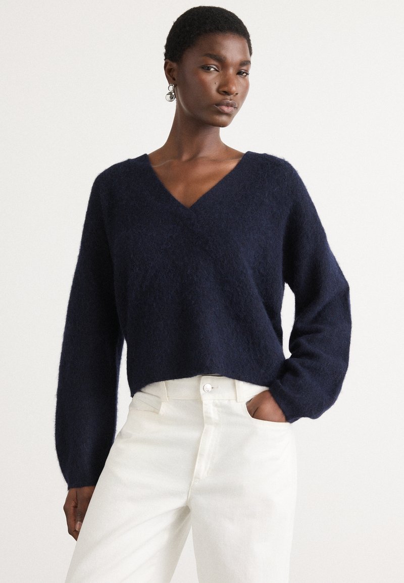 Navyblauwe, korte trui van zacht, aaibaar materiaal met een V-hals en lange, losse mouwen, gecombineerd met witte, high-waisted broek.