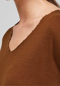 Brun stickad tröja med V-ringning, prydd med små metallnaglar längs kanten. Slät textur och åtsittande design.