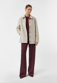 Woman standing wearing beige buttoned coat, burgundy sweater, wide-leg burgundy trousers, and matching pointed shoes against white background.