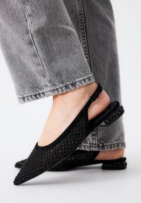 Primer plano de pies que llevan unas bailarinas negras de malla con pequeños adornos de piedras en los tacones, combinadas con unos jeans grises.