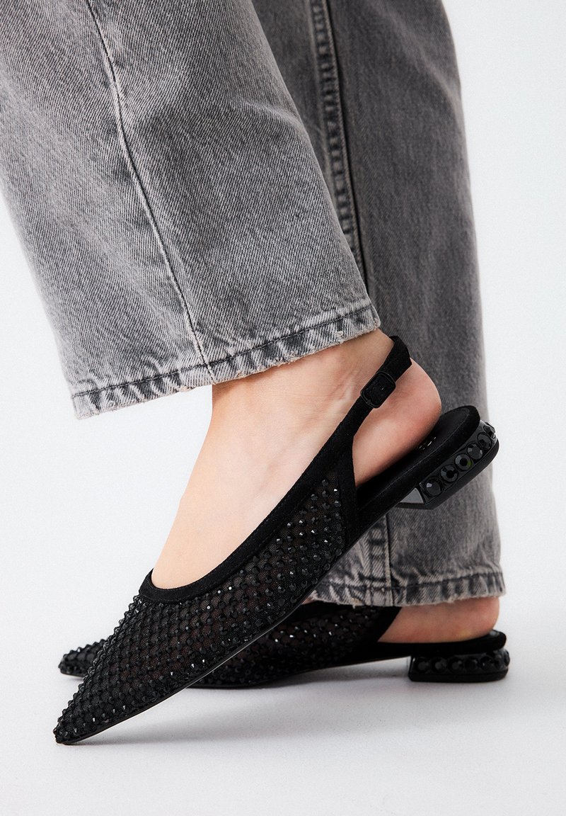 Primer plano de pies que llevan unas bailarinas negras de malla con pequeños adornos de piedras en los tacones, combinadas con unos jeans grises.