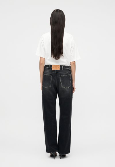 Άτομο με μακριά ίσια μαλλιά στέκεται γυρισμένο πλάτη, φορώντας λευκό t-shirt, μαύρα τζιν παντελόνια φαρδιάς γραμμής με τσέπες πίσω και μαύρες ψηλοτάκουνες γόβες.