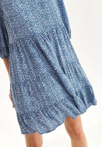 Kurzes, blaues Blumenkleid aus leichtem Stoff. Es hat einen gerafften Saum, eine lockere Passform und Dreiviertelärmel, die mit kleinen weißen Blumen gemustert sind.