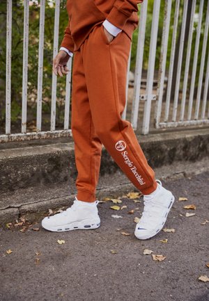 Osoba ubrana w rdzawe spodnie dresowe z logo „Sergio Tacchini” i białe buty sportowe Nike, stojąca na chodniku pokrytym liśćmi przy metalowym ogrodzeniu.