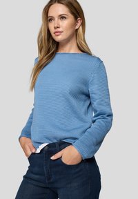 Junge Frau mit langen Haaren, die einen hellblauen strukturierten Pullover und dunkle blaue Jeans trägt, steht mit den Händen in den Taschen vor einem neutralen Hintergrund.