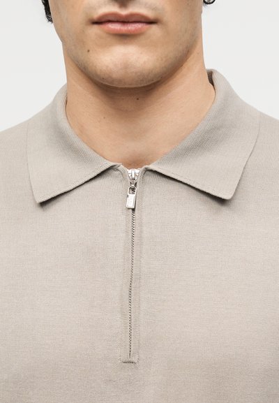 Άνδρας με μπεζ πλεκτό μπλουζάκι με γιακά και με φερμουάρ στο μπροστινό μέρος μέχρι τη μέση, που δείχνει τα χείλη, το πηγούνι και τον λαιμό σε απλό φόντο.