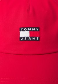 Punainen hattu kestävästä kankaasta, jossa on brodeerattu musta-valkoinen etiketti, jossa lukee "TOMMY JEANS" sekä värillinen logosuunnittelu.