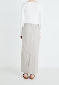 Frau trägt ein weißes Langarmshirt, einen beigefarbenen knöchellangen Rock und braune flache Sandalen, steht und blickt auf eine weiße Wand.