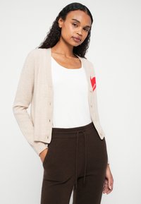 Beżowy kardigan wykonany z miękkiego materiału, z czerwonym sercem na piersi, zapinany na guziki, z długimi rękawami. W komplecie z białą bluzką.