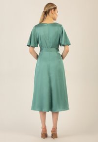 Un vestito verde di raso con maniche a farfalla corte, vita aderente e gonna di media lunghezza. Texture liscia e lucentezza sottile in tutto il capo.