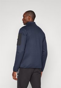 Námornícky fleecový jacket s vysokým golierom, textúrovanou tkaninou a čiernou nášivkou na ľavom rukáve. Navrhnutý pre pohodlie a teplo.