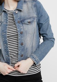 Ljusblå denimjacka med två bröstfickor, metalldrivna knappar och en figurnära design. Under jackan en svart- och vitrandig skjorta.