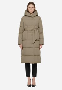 Piumino color marrone chiaro con cappuccio, vita stretta da una cintura e design trapuntato. Maniche lunghe e lunghezza fino al ginocchio, abbinato a pantaloni neri e tacchi.