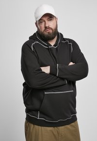 Felpa nera con cuciture grigie, tasca frontale e cappuccio con cordino. Indossata con pantaloni beige e un berretto bianco. Morbido tessuto.