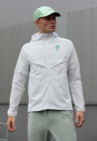 Muž v bielej zipsovej bunde s zeleným logom, svetlozelených nohaviciach a svetlozelenom klobúku, stojaci vonku na šedom pozadí.