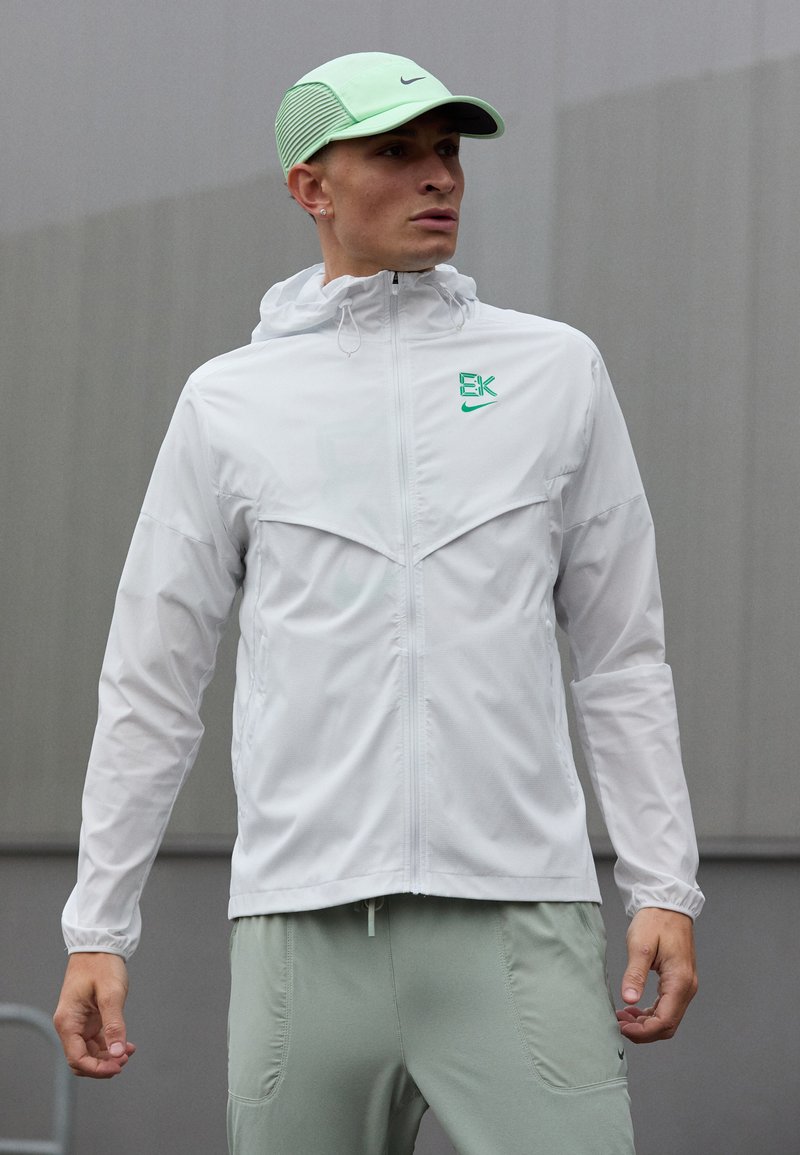 Muž v bielej zipsovej bunde s zeleným logom, svetlozelených nohaviciach a svetlozelenom klobúku, stojaci vonku na šedom pozadí.