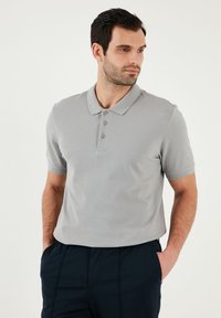 Šedé polo tričko s krátkým rukávem, klasickým límcem, třemi knoflíky, strukturovanou tkaninou a uvolněným střihem, spárované s tmavě námořnickými kalhotami.