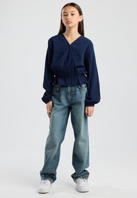 Navy blue textured blouse with a wrap design and elastic waistband, paired with light blue straight-leg jeans and white sneakers.