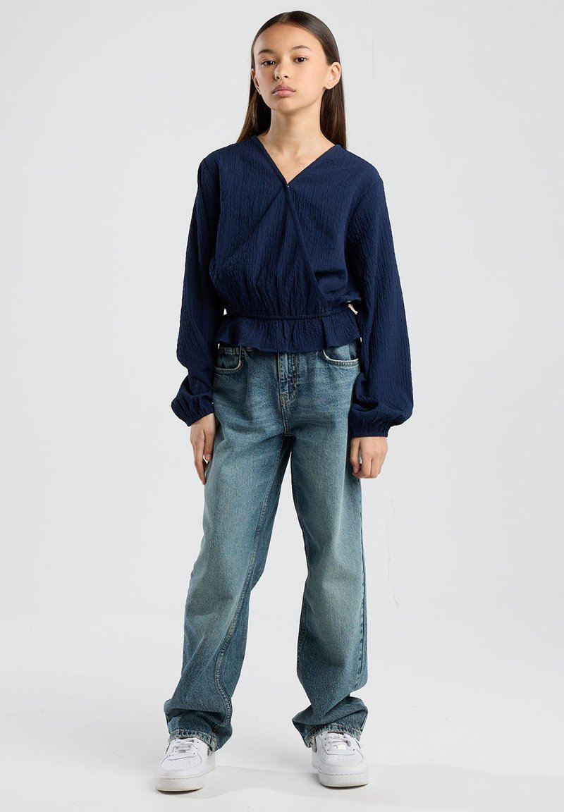 Navy blue textured blouse with a wrap design and elastic waistband, paired with light blue straight-leg jeans and white sneakers.