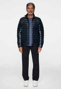 Pehmeä, tummansininen takki, jossa on kiiltävä pinta, etuvetoketju ja kohotettu kaulus. Päälläsininen t-paita, yhdistettynä tummiin housuihin ja valkoisiin kenkiin.