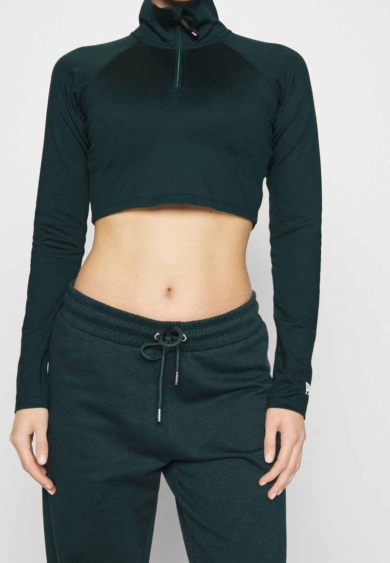 Tmavě zelený, zkrácený sportovní top s vysokým límcem a dlouhými rukávy. Spárovaný s odpovídajícími tmavě zelenými tepláky, které mají elastický pas a tkaničku.